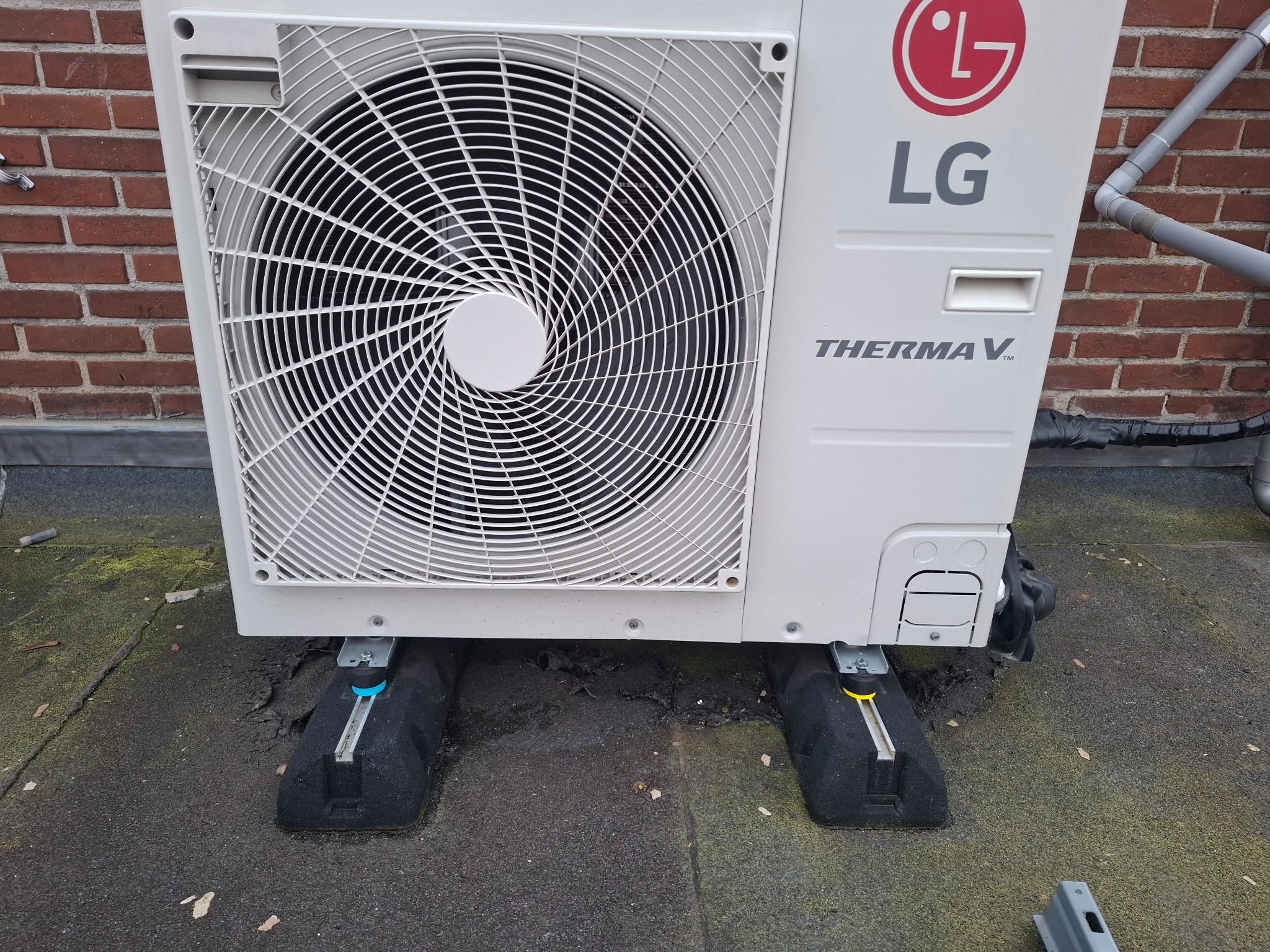Biała jednostka zewnętrzna pompy ciepła LG Therma V, zamontowana na czarnych podkładkach antywibracyjnych na dachu. Widoczna ceglana ściana w tle.