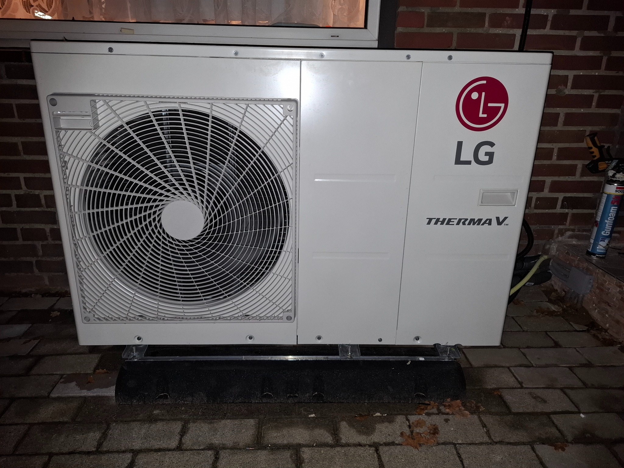 Biała pompa ciepła LG Therma V, zamontowana na zewnątrz budynku z cegły, widoczna kratka wentylatora i logo producenta. Obok narzędzia montażowe.