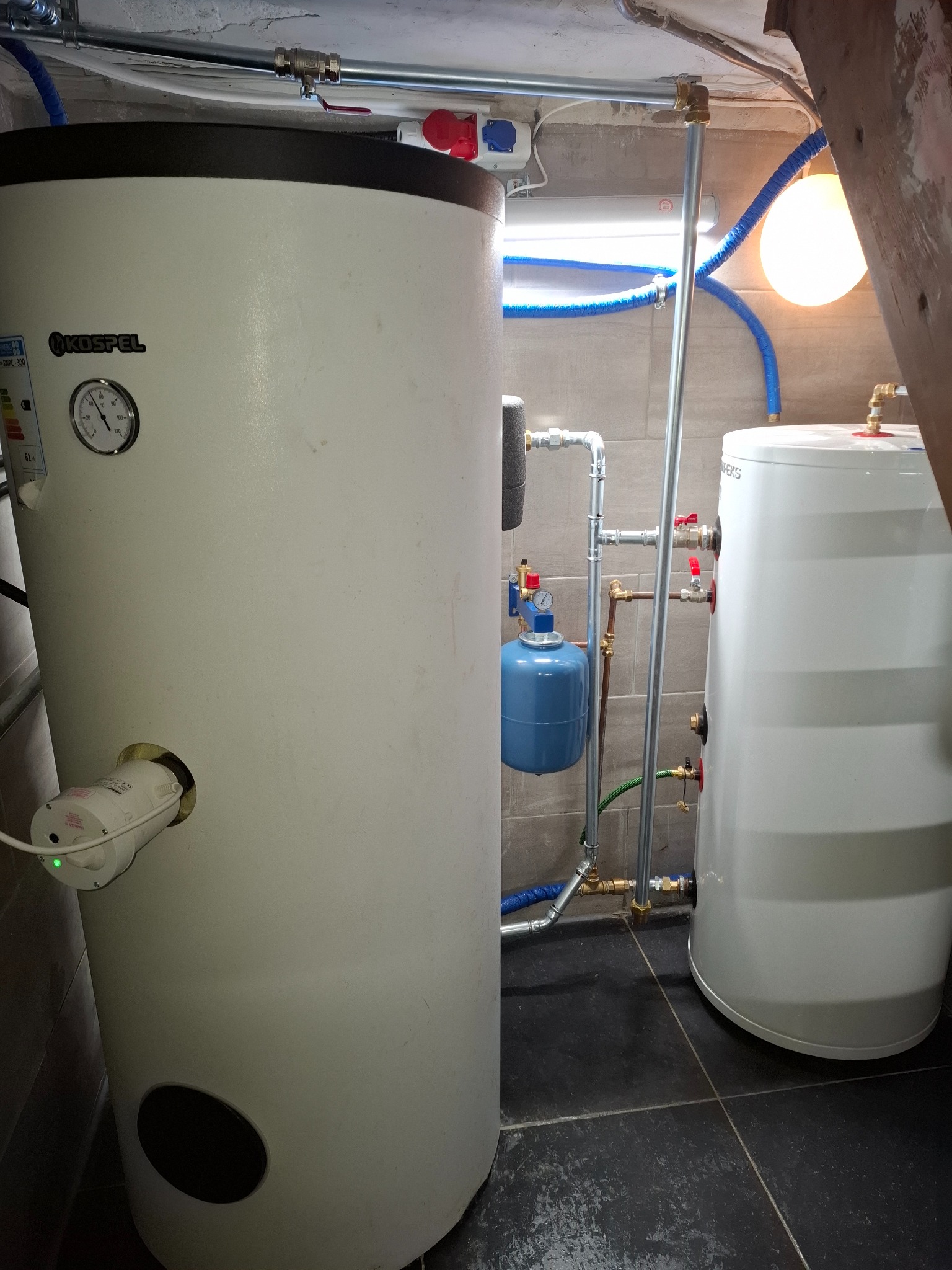 Dwa białe zasobniki ciepłej wody użytkowej Kospel z podłączeniami hydraulicznymi i elektrycznymi w kotłowni z szarą glazurą. Widoczne rury i osprzęt.