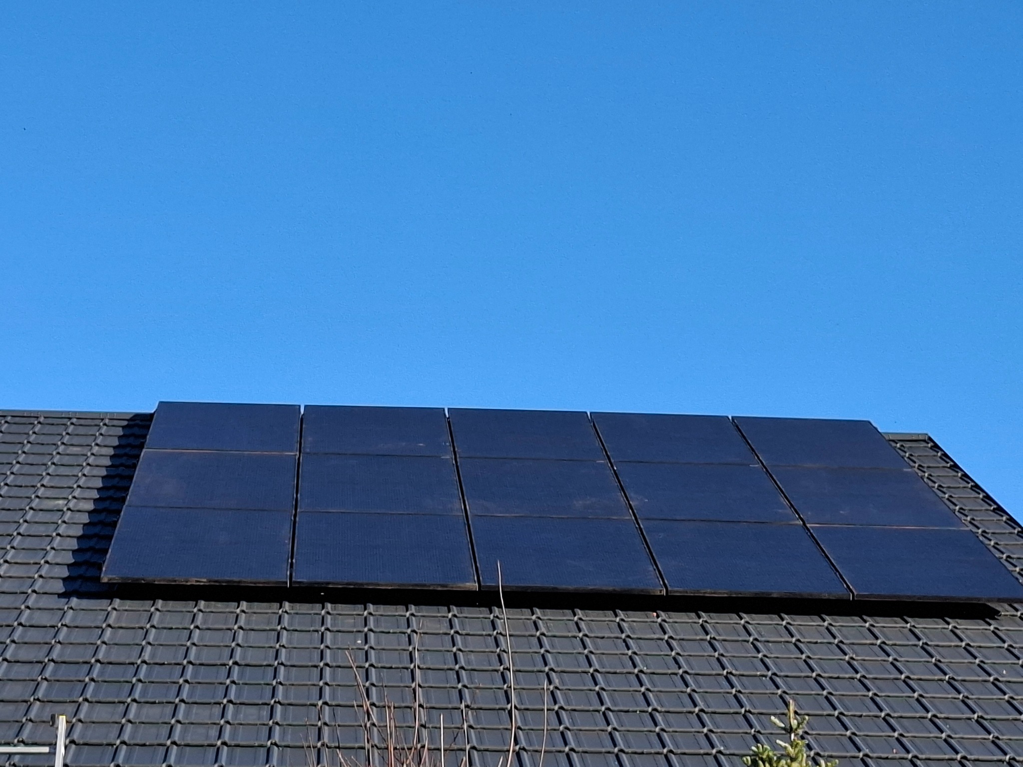Panele fotowoltaiczne na dachu domu z ciemnej dachówki, widoczne z dołu pod kątem, na tle błękitnego nieba. Widoczny fragment drabiny.
