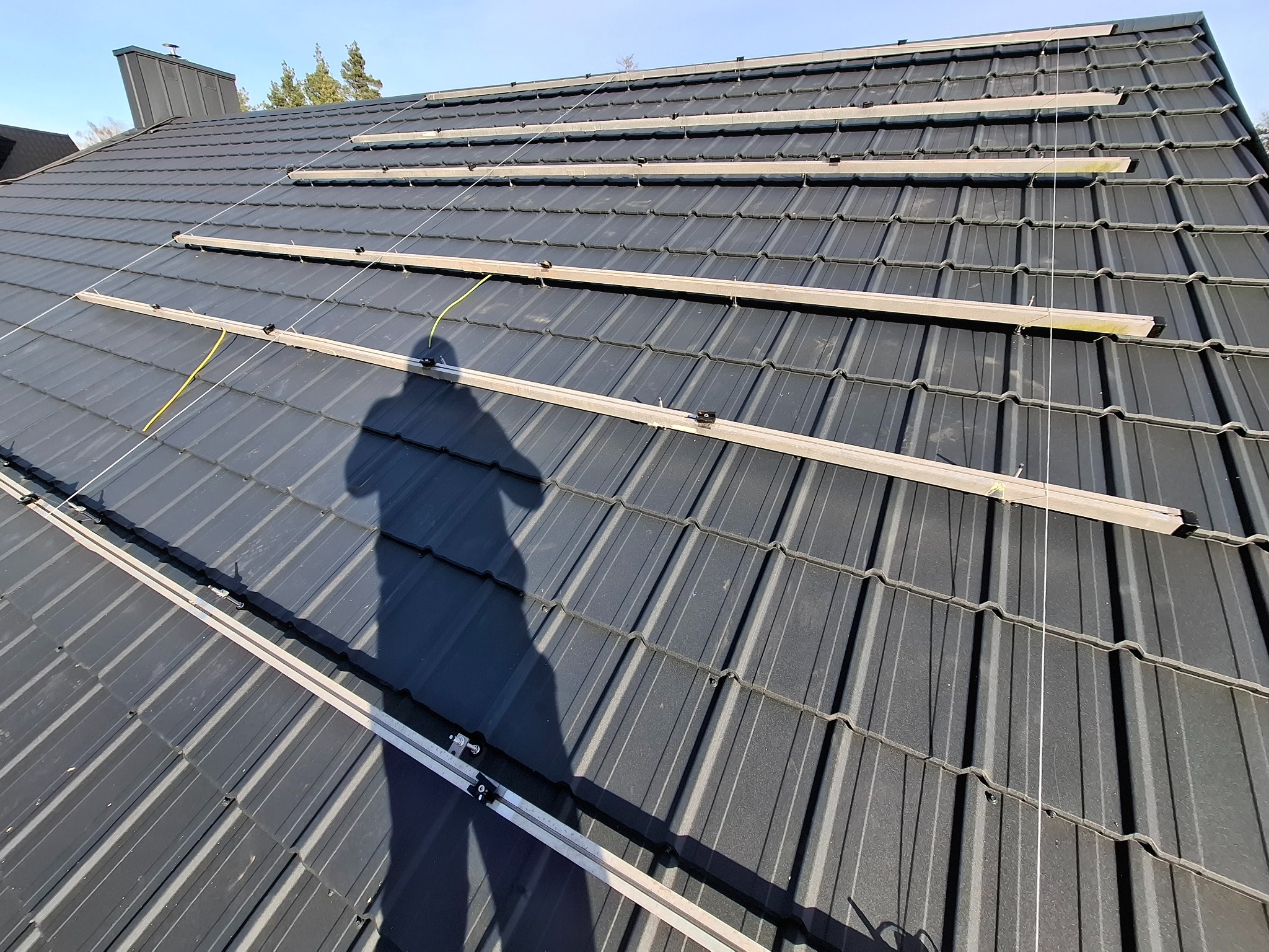 Montaż szyn pod panele fotowoltaiczne na grafitowym dachu z blachodachówki, cień osoby w lewym dolnym rogu. Widok z góry, słoneczny dzień.