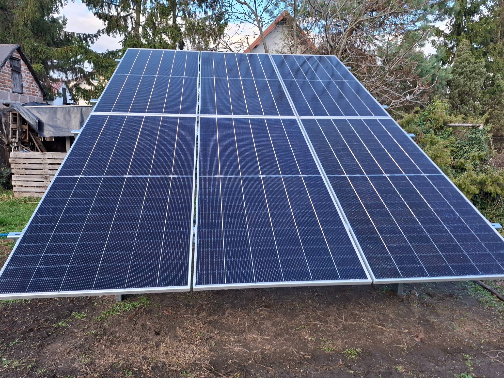 Ustawione panele fotowoltaiczne na ziemi, widoczne z bliska, z domem i zielenią w tle. Układ paneli 3x2, srebrne ramy, ciemnoniebieskie ogniwa.