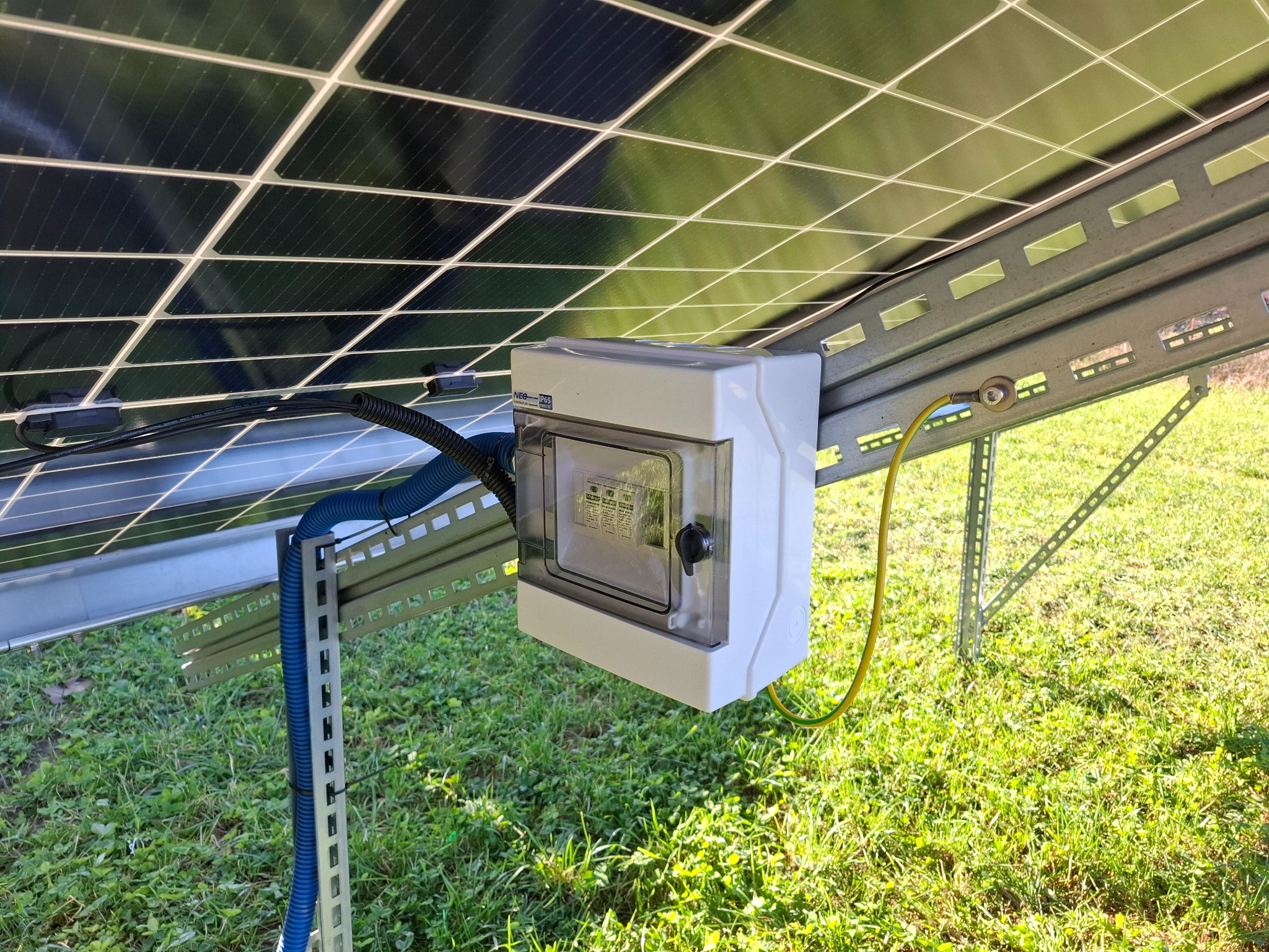 Panel fotowoltaiczny od spodu z puszką przyłączeniową i kablem uziemiającym na tle zielonej trawy. Widoczne elementy montażowe i instalacyjne.