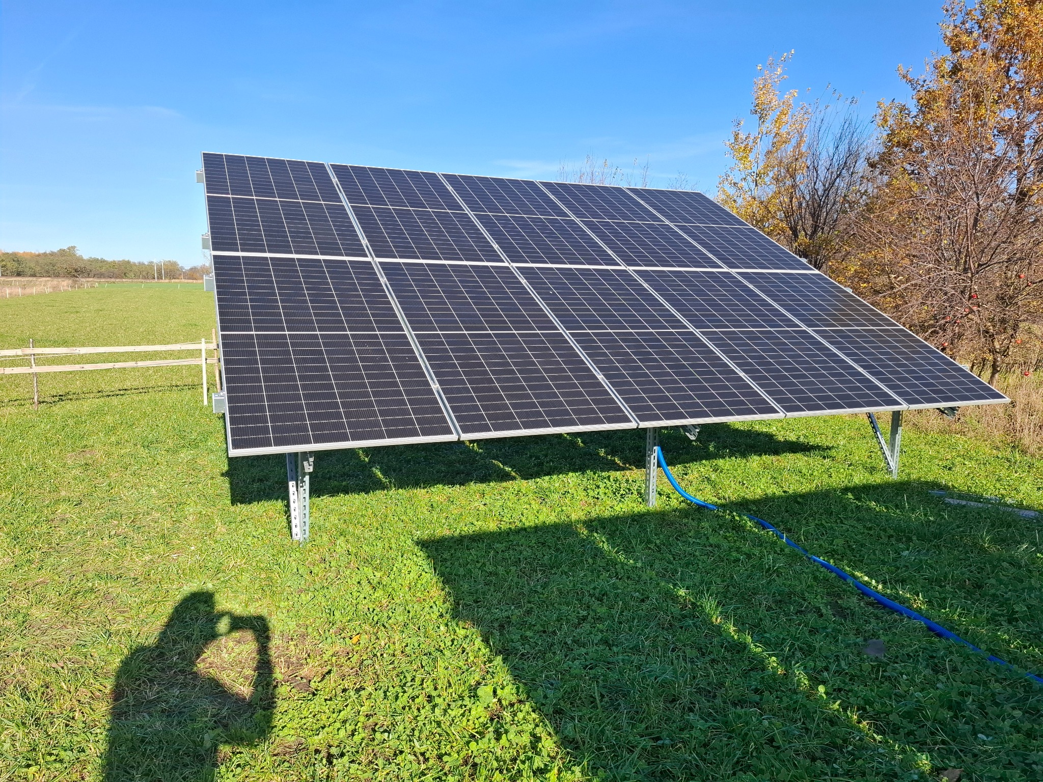 Panel słoneczny na trawie w słoneczny dzień. Widoczny cień osoby robiącej zdjęcie. Błękitny przewód leży na trawie. W tle drzewa i niebo.