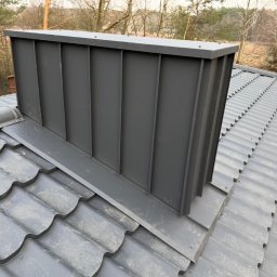 Andrzej Swojak - Szary komin z blachy na dachu z szarej dachówki karpiówki, widok z góry, drzewa w tle. Komin posiada pionowe przetłoczenia i płaskie wykończenie.