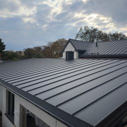 Andrzej Swojak - Nowoczesny, grafitowy dach z blachy na domu jednorodzinnym, widok z góry, z widocznymi detalami obróbek blacharskich i rynien. Pochmurne niebo w tle.