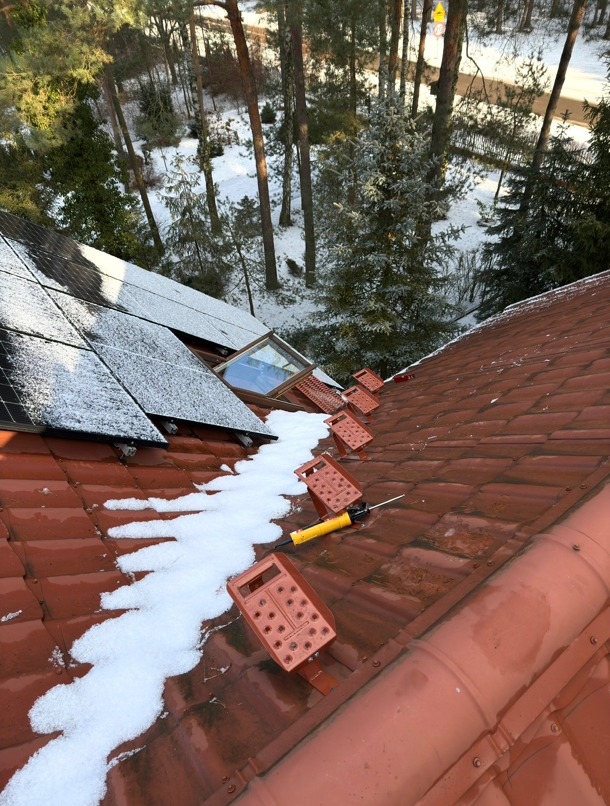 Czerwony dach z panelami słonecznymi i oknem dachowym, pokryty śniegiem. Widoczne narzędzia i elementy montażowe. W tle ośnieżony las.