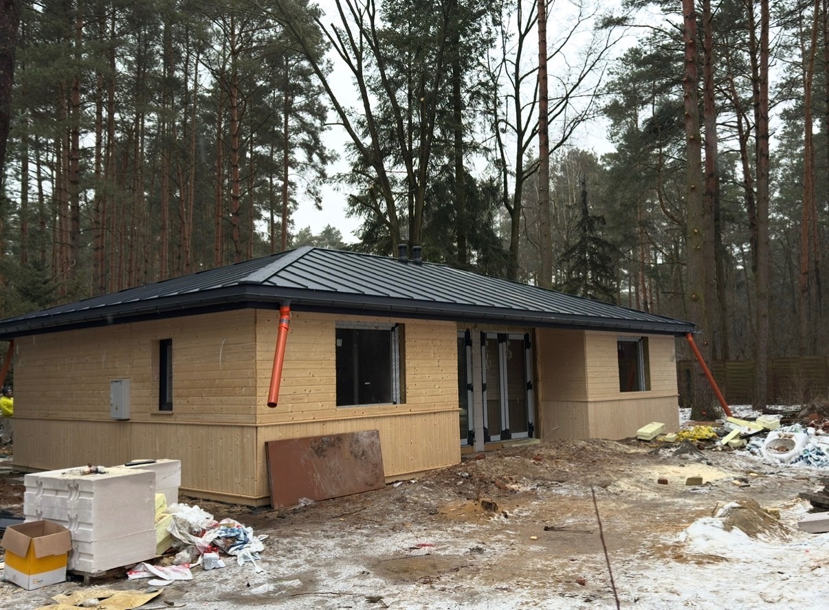 Drewniany dom z ciemnym dachem na tle lasu. Widoczne rynny i elementy wykończeniowe. Teren wokół domu w trakcie prac budowlanych, z widocznymi materiałami.