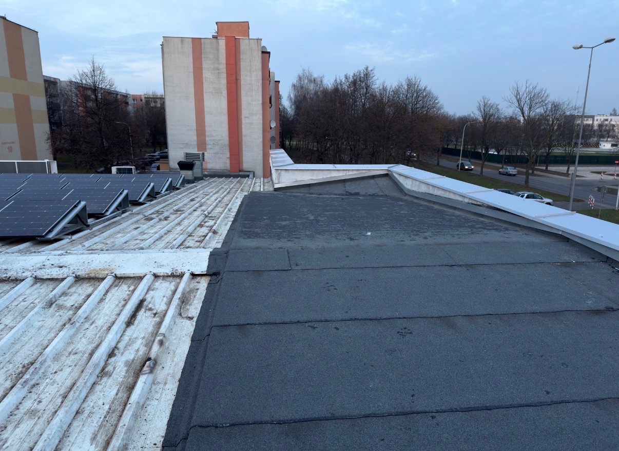 Płaski dach budynku z panelami słonecznymi i pokryciem z papy bitumicznej. Widoczne budynki mieszkalne w tle i fragment ulicy.