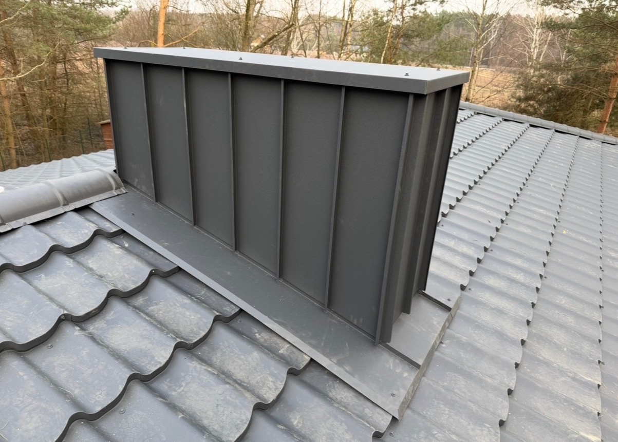 Szary komin z blachy na dachu z szarej dachówki karpiówki, widok z góry, drzewa w tle. Komin posiada pionowe przetłoczenia i płaskie wykończenie.