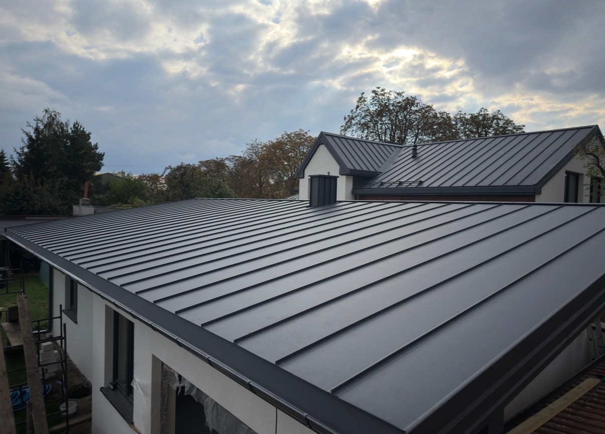 Nowoczesny, grafitowy dach z blachy na domu jednorodzinnym, widok z góry, z widocznymi detalami obróbek blacharskich i rynien. Pochmurne niebo w tle.