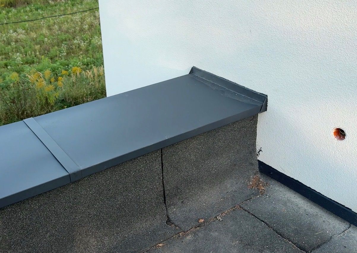 Szczegółowe ujęcie obróbki blacharskiej na dachu z papy, z widocznym połączeniem z elewacją budynku. Nowoczesne wykończenie, precyzyjne łączenia blachy.