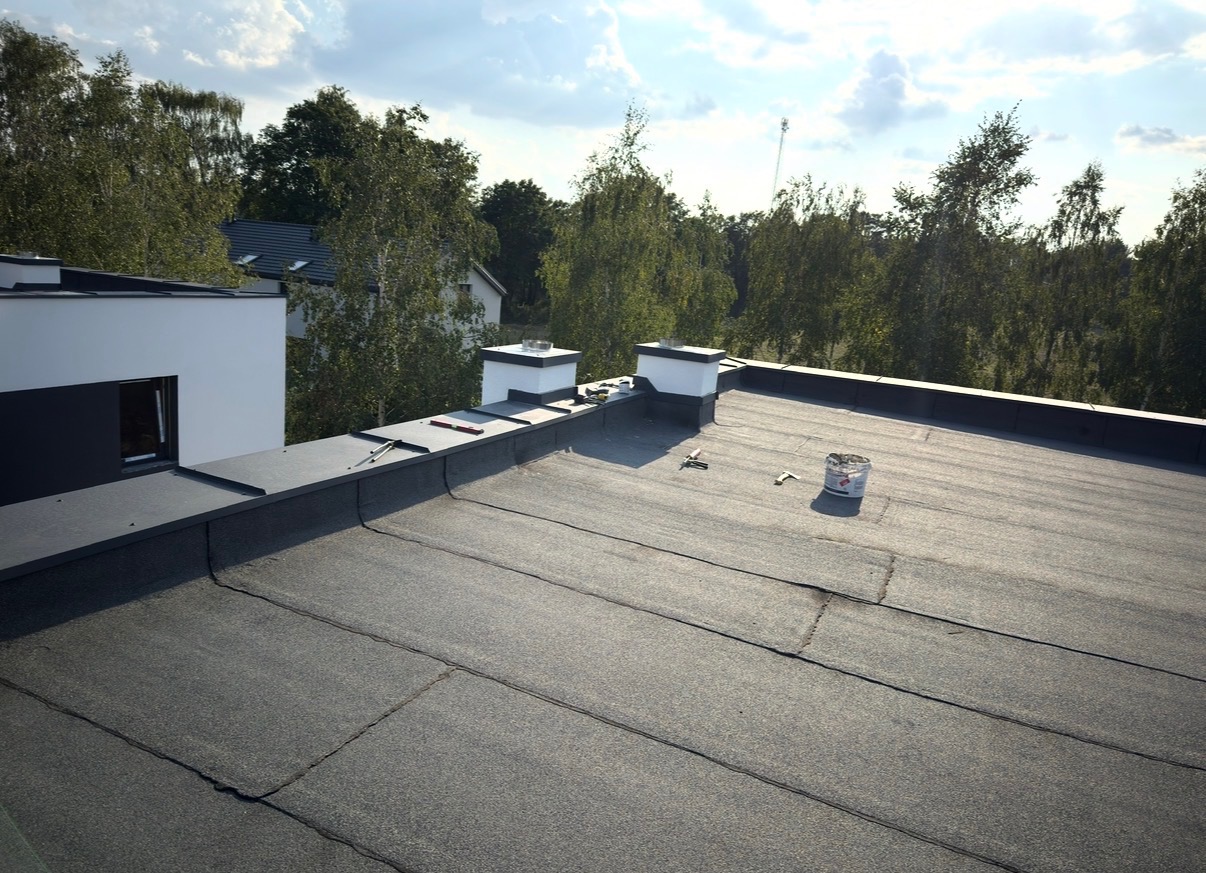 Dach pokryty papą termozgrzewalną z detalami obróbek blacharskich przy kominach i krawędziach. Widoczne narzędzia i puszka farby. Tło: zieleń drzew i fragment budynku.