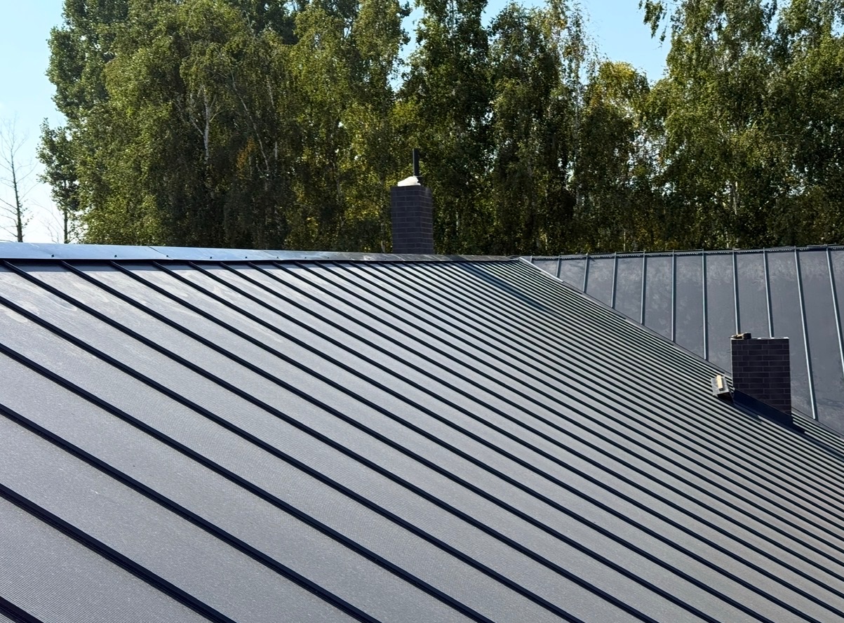 Szary dach z blachy na tle zielonych drzew i błękitnego nieba. Widoczne dwa kominy z cegły. Ujęcie z dołu, podkreślające regularność i nowoczesny wygląd pokrycia.