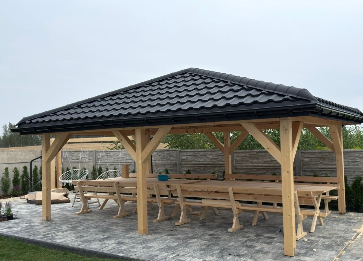 Zadaszona drewniana altana z czarnym dachem z blachodachówki, widok z zewnątrz na stół z ławkami, szare płytki na podłodze i huśtawka w tle.
