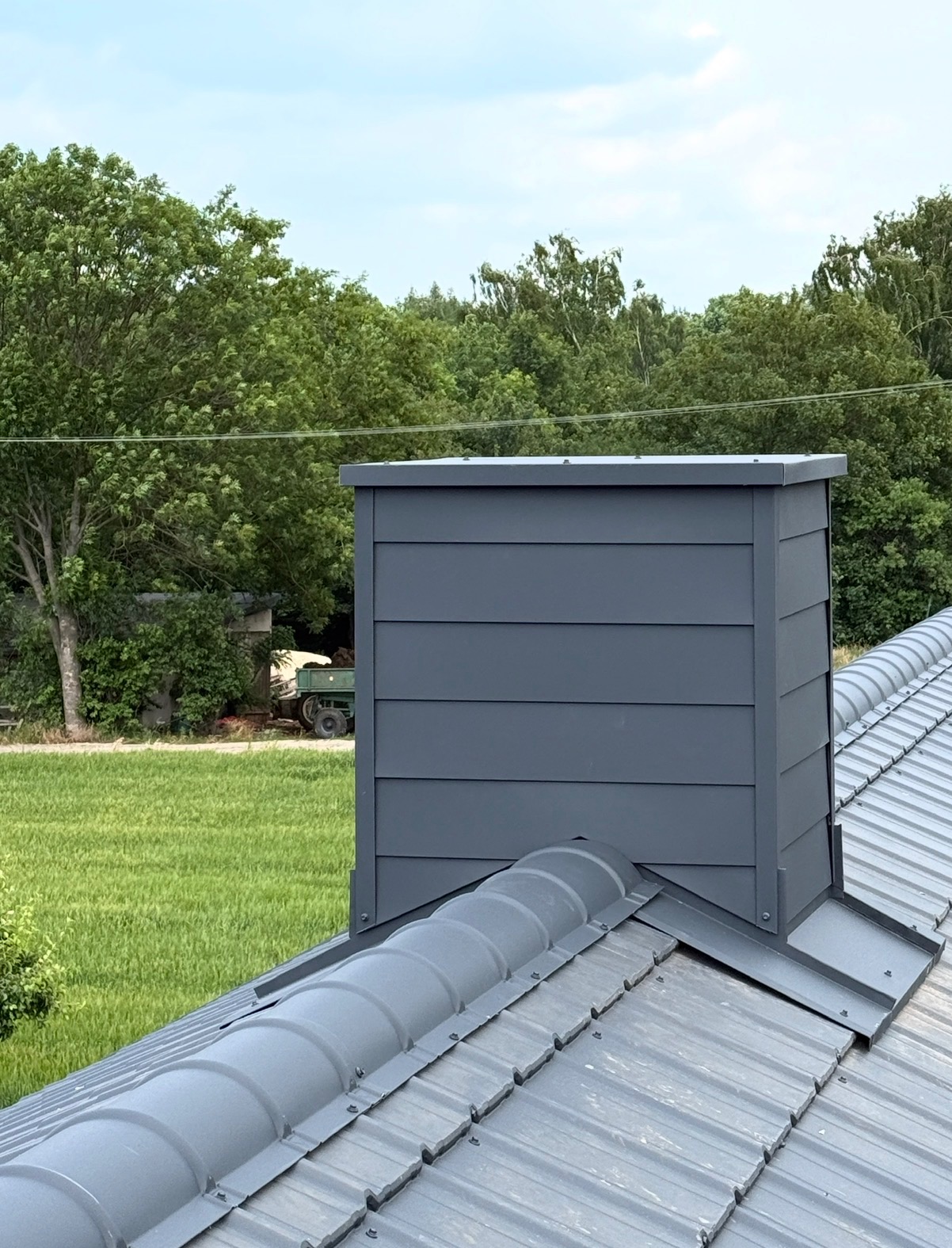 Szary komin z blachy na dachu z blachy trapezowej, na tle zieleni drzew i łąki. Komin wykończony panelami, widoczne łączenia blacharskie.