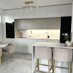 invero studio - Nowoczesna kuchnia z białymi frontami, marmurowym blatem i złotymi akcentami, oświetlona lampą sufitową z kulistymi kloszami. Widoczne krzesła barowe z beżową tapicerką.