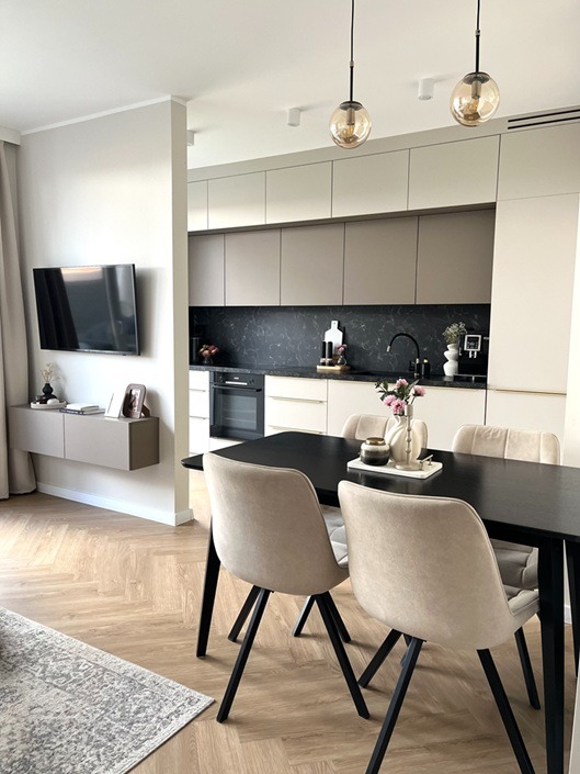 Nowoczesna kuchnia z jadalnią: beżowe szafki, czarny blat, stół z krzesłami. Parkiet w jodełkę, wiszące lampy. Minimalistyczny design, jasne wnętrze.