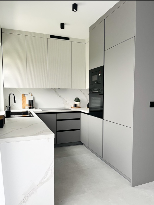 Nowoczesna, narożna kuchnia w odcieniach szarości i bieli z marmurowym blatem i minimalistycznym oświetleniem punktowym. Fronty górnych szafek z subtelnym, pionowym frezem.