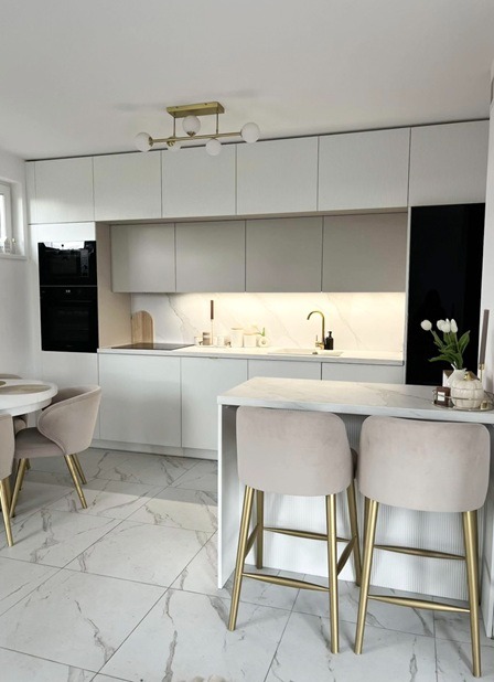 Nowoczesna kuchnia z białymi frontami, marmurowym blatem i złotymi akcentami, oświetlona lampą sufitową z kulistymi kloszami. Widoczne krzesła barowe z beżową tapicerką.