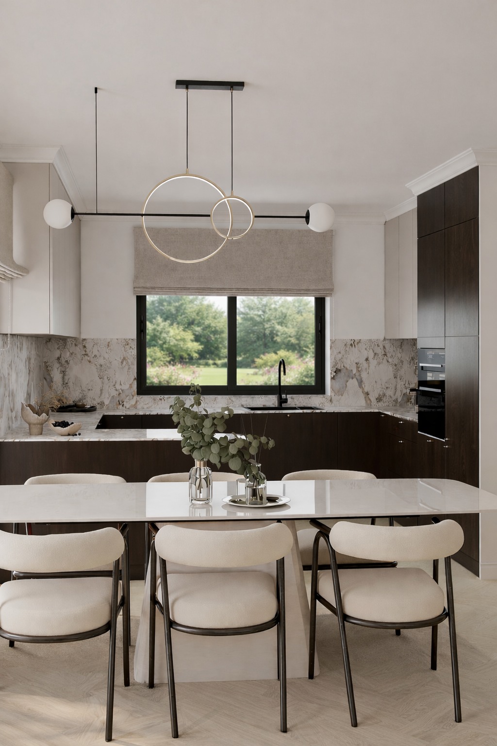 Nowoczesna kuchnia z marmurowym blatem i ciemnymi szafkami, oświetlona lampą z okręgów. Widok na ogród przez okno z roletą rzymską. Stół z krzesłami w neutralnych barwach.