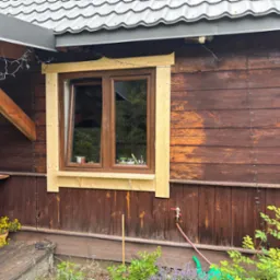 Drewniany dom z oknem w trakcie montażu obramowania. Widoczne surowe drewno i dekoracyjne lampki. Częściowo odnowiona elewacja, z widocznymi śladami wcześniejszej farby.