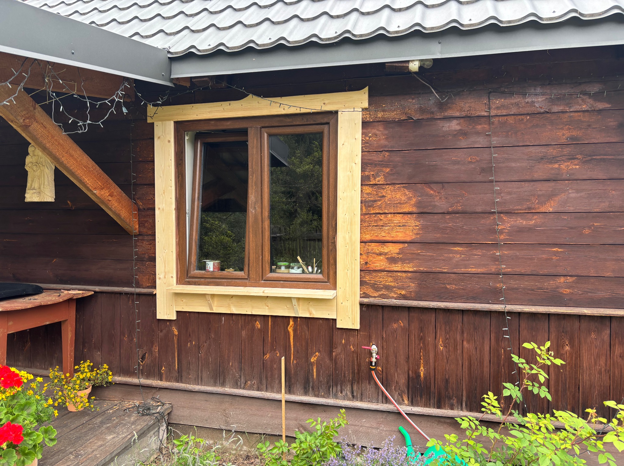 Drewniany dom z oknem w trakcie montażu obramowania. Widoczne surowe drewno i dekoracyjne lampki. Częściowo odnowiona elewacja, z widocznymi śladami wcześniejszej farby.