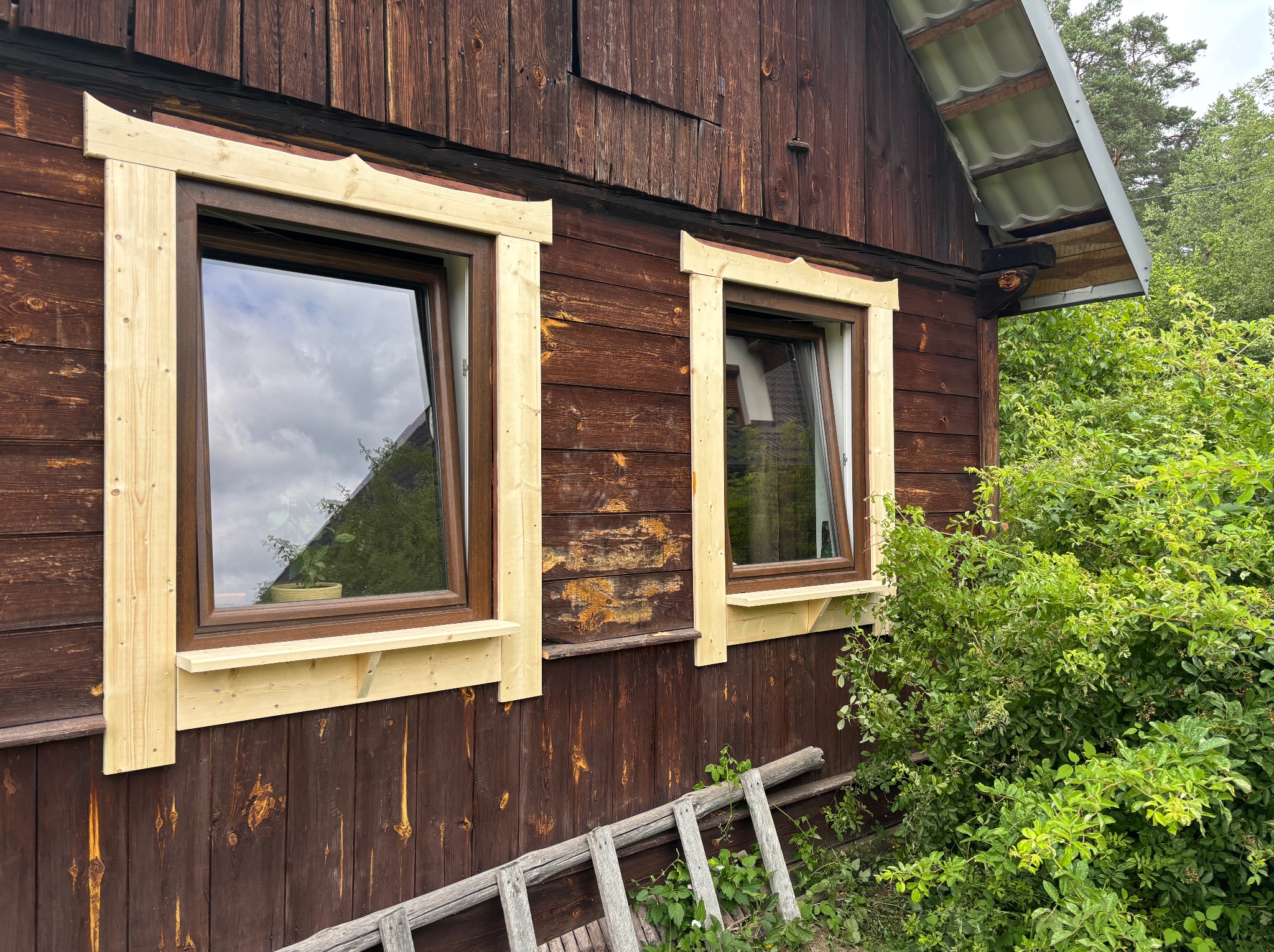 Brunatna drewniana elewacja z dwoma oknami w brązowych ramach, ozdobionymi jasnym, surowym drewnem. W tle zieleń i fragment dachu.