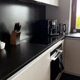 Nowoczesna kuchnia z czarnym blatem, indukcyjną płytą i ekspresem do kawy. Minimalistyczny design, białe szafki, drewniane akcenty. Widok na okno z zielenią.