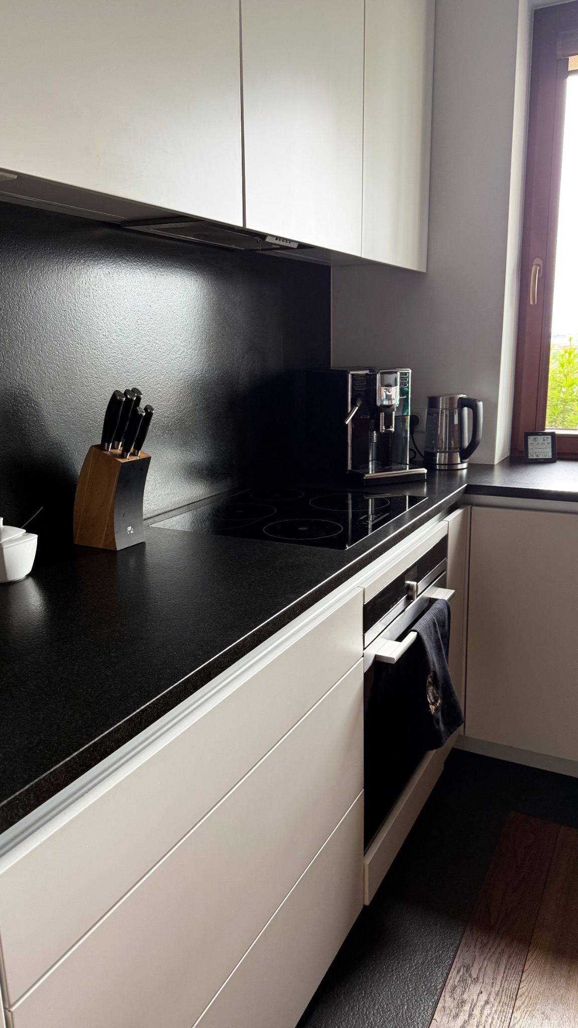 Nowoczesna kuchnia z czarnym blatem, indukcyjną płytą i ekspresem do kawy. Minimalistyczny design, białe szafki, drewniane akcenty. Widok na okno z zielenią.