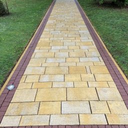 PanCzyścik Czyszczenie Kostki Brukowej - Ułożona z beżowo-żółtej kostki brukowej ścieżka z bordową obwódką, oświetlona punktowo, prowadząca przez trawnik w kierunku drewnianej altany.