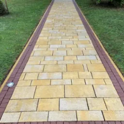 Ułożona z beżowo-żółtej kostki brukowej ścieżka z bordową obwódką, oświetlona punktowo, prowadząca przez trawnik w kierunku drewnianej altany.