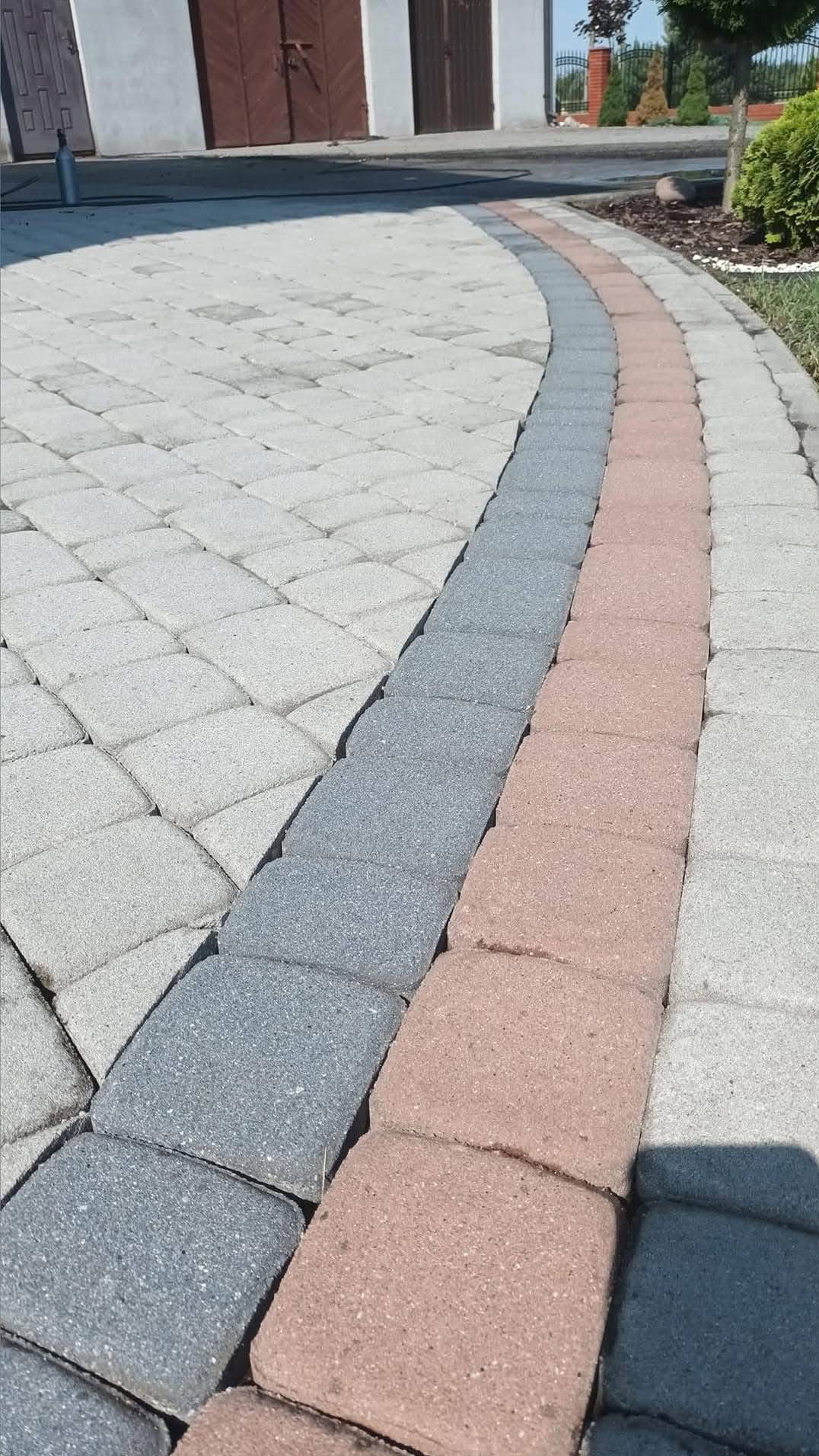 Ułożona kostka brukowa w trzech odcieniach szarości, tworząca łukowaty wzór na podjeździe przed budynkiem z drewnianymi wrotami.