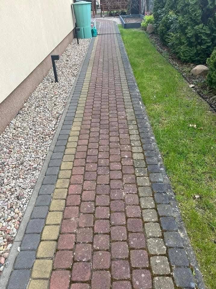 Chodnik z kostki brukowej w odcieniach czerwieni, żółci i szarości, częściowo porośnięty mchem, przylegający do ściany budynku z jasnymi kamykami i trawnika z zieloną trawą.