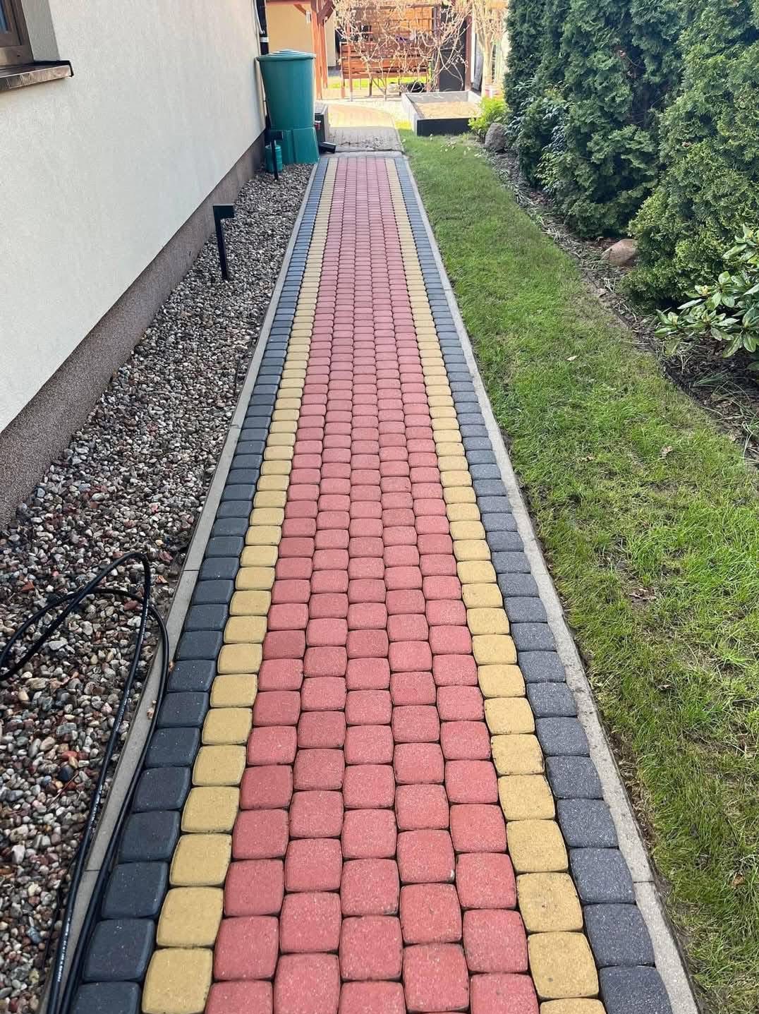 Ścieżka z kolorowej kostki brukowej (czerwonej, żółtej i szarej) prowadząca wzdłuż ściany domu i trawnika, z widocznym zbiornikiem na deszczówkę w tle.
