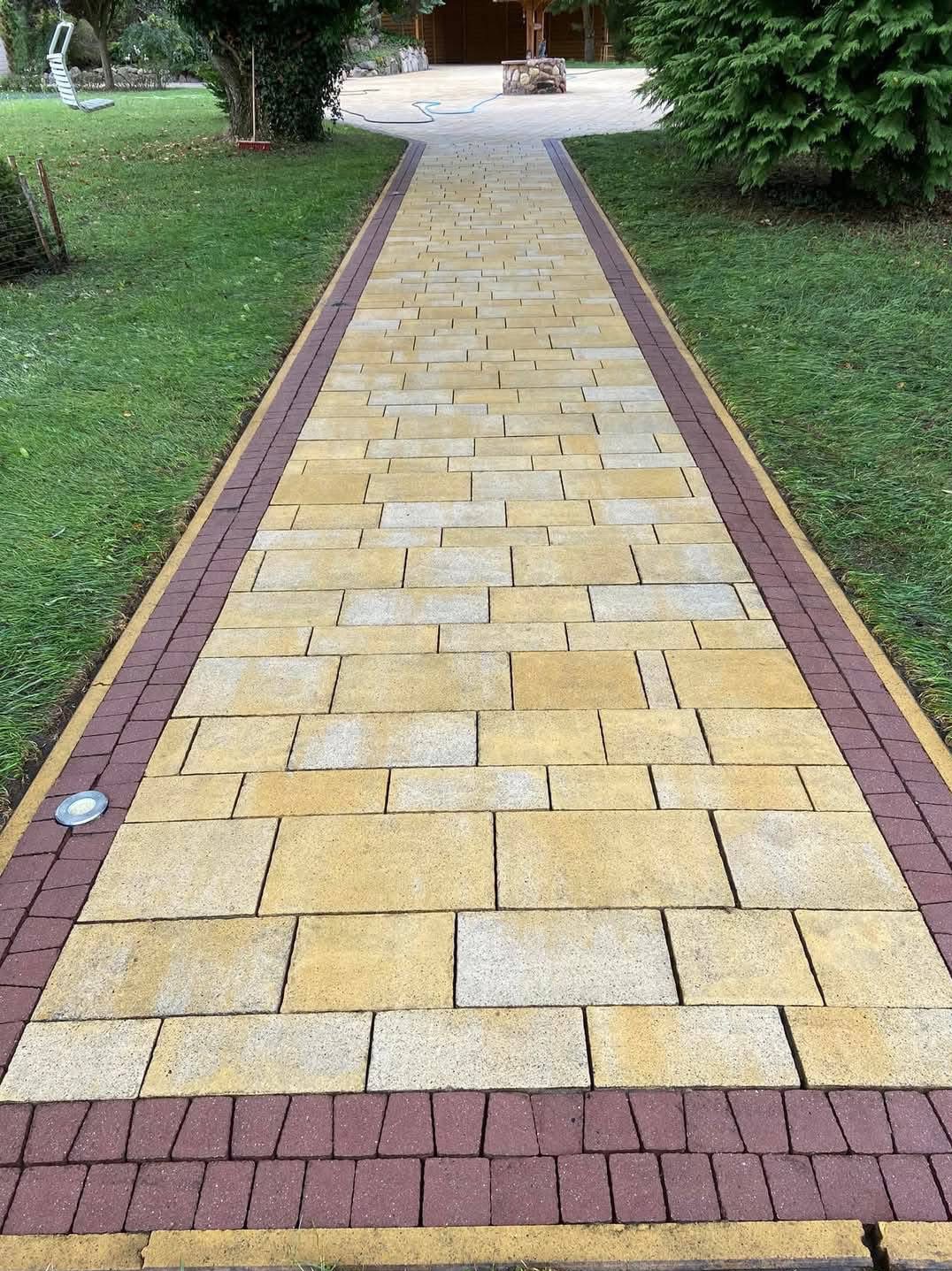 Ułożona z beżowo-żółtej kostki brukowej ścieżka z bordową obwódką, oświetlona punktowo, prowadząca przez trawnik w kierunku drewnianej altany.