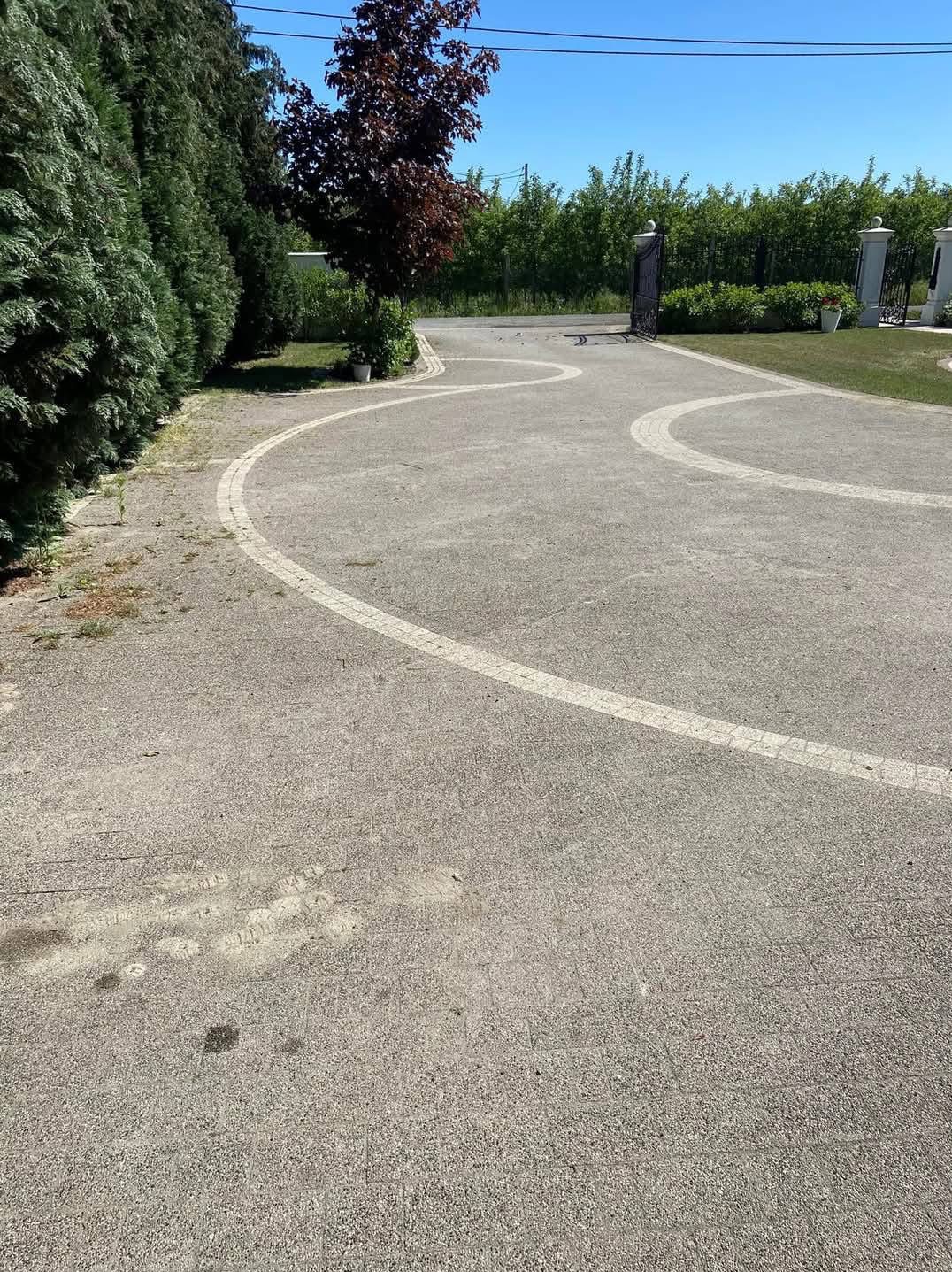 Szeroki podjazd z kostki brukowej z dekoracyjnymi, jasnymi obwódkami w kształcie wijących się linii, otoczony zielenią i prowadzący do bramy wjazdowej.