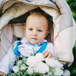 Adrian Szymkowiak CREACTIVE - Niebieskooki niemowlak w wózku, ubrany w białą koszulę, błękitną muchę i szelki, trzyma bukiet białych kwiatów.