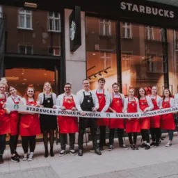 Zespół pracowników Starbucks w Chodzieży pozuje przed wejściem do kawiarni, trzymając baner z nazwą i adresem lokalu.