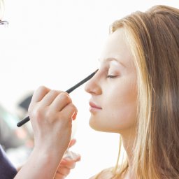 Adrian Szymkowiak CREACTIVE - Wizażystka nakłada pędzlem cień na powiekę klientki z blond włosami, ujęcie z profilu, jasne tło.