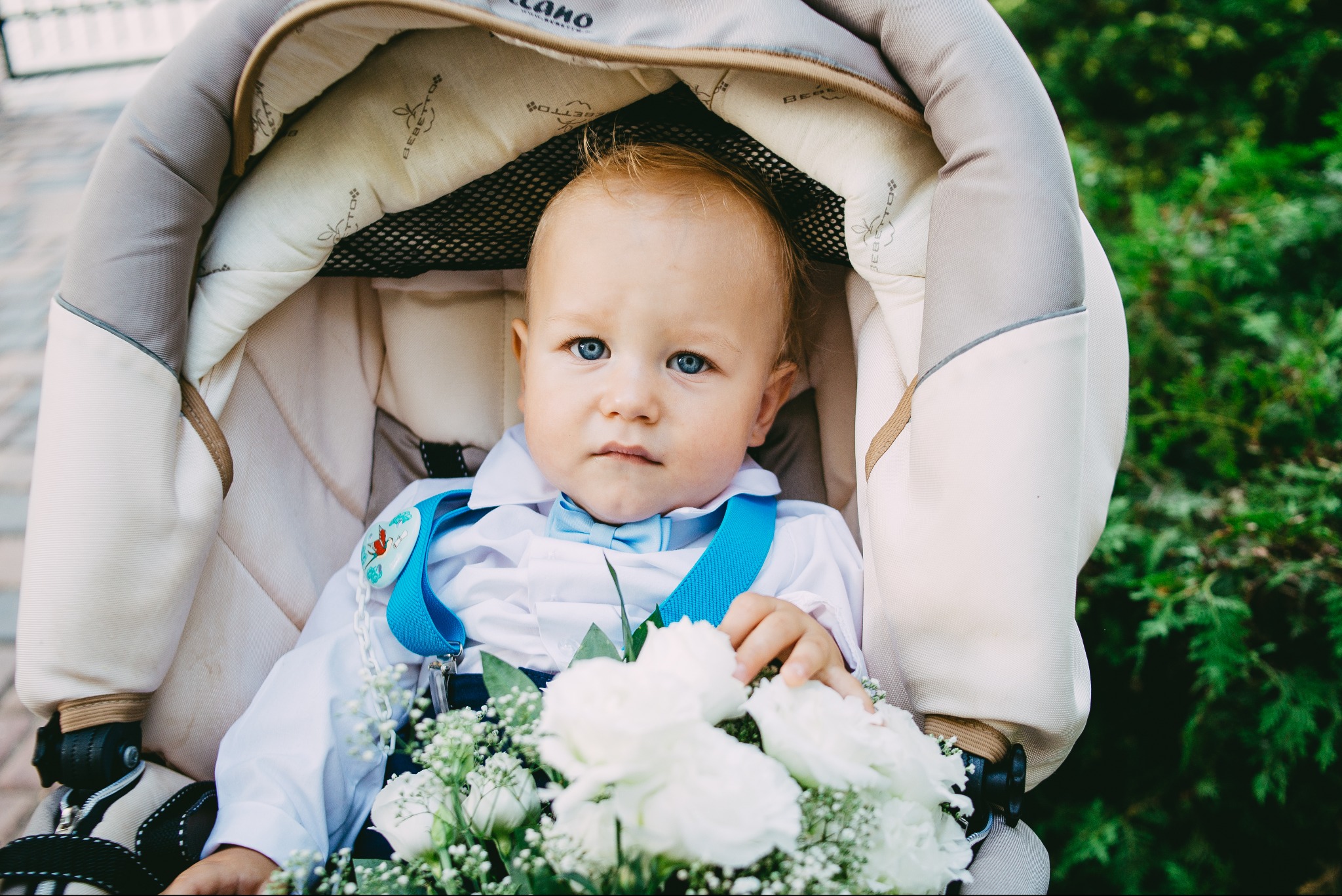 Niebieskooki niemowlak w wózku, ubrany w białą koszulę, błękitną muchę i szelki, trzyma bukiet białych kwiatów.