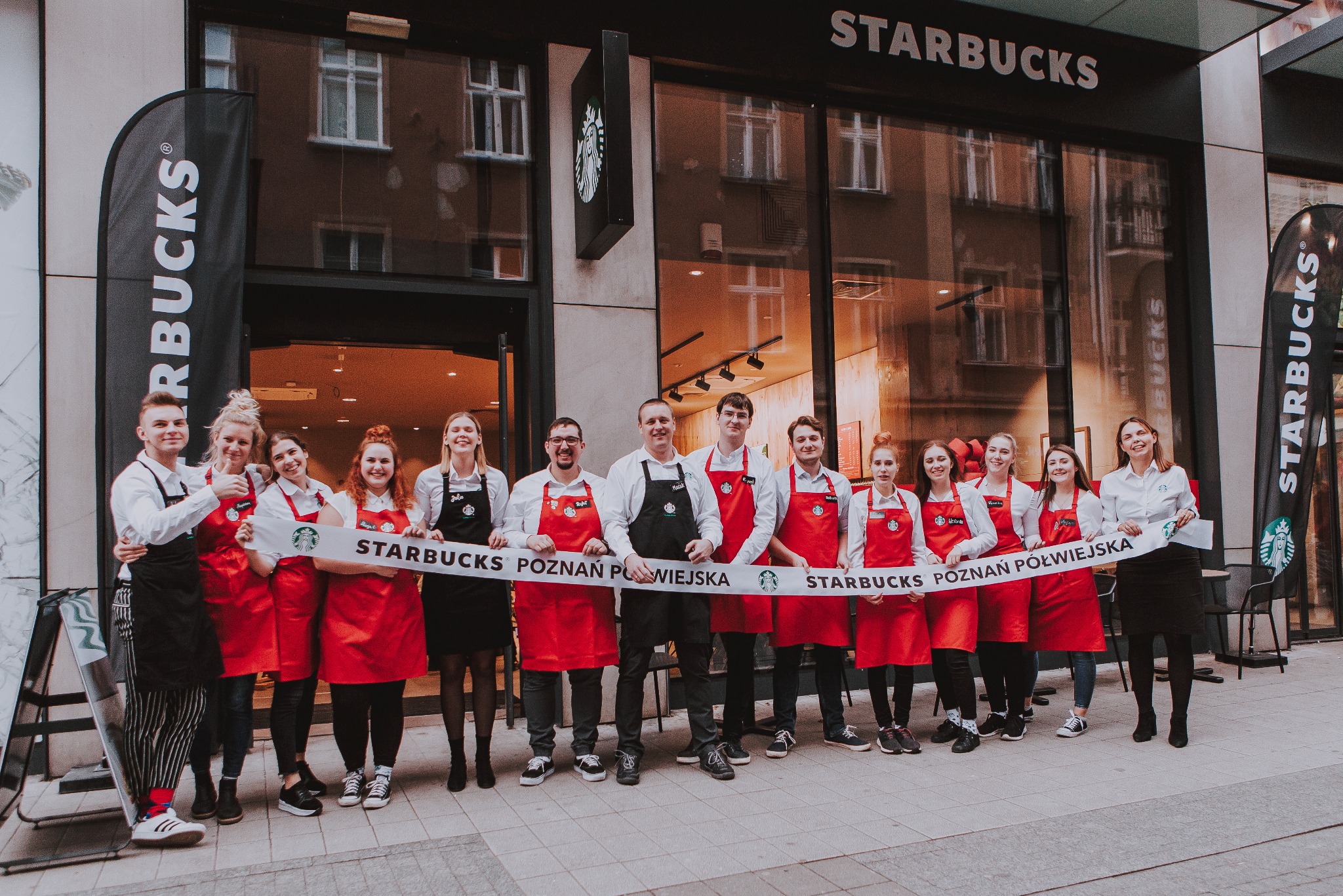 Zespół pracowników Starbucks w Chodzieży pozuje przed wejściem do kawiarni, trzymając baner z nazwą i adresem lokalu.