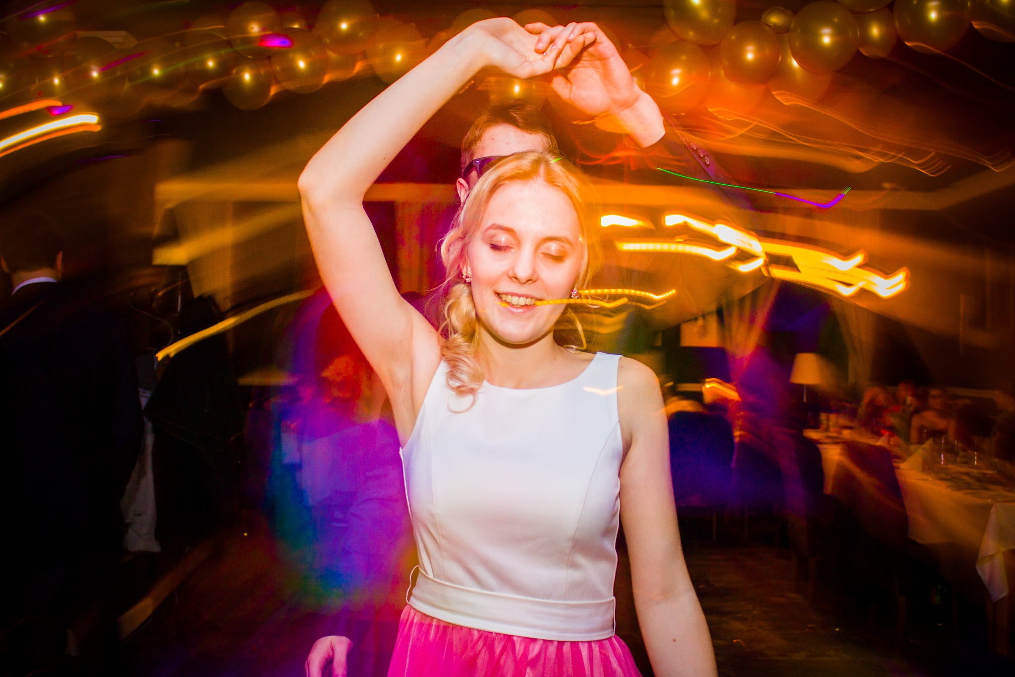 Uśmiechnięta blondynka w białej sukience i różowej spódnicy tańczy z podniesionymi rękami na rozświetlonym parkiecie podczas imprezy, trzymając w ustach świecące pałeczki.