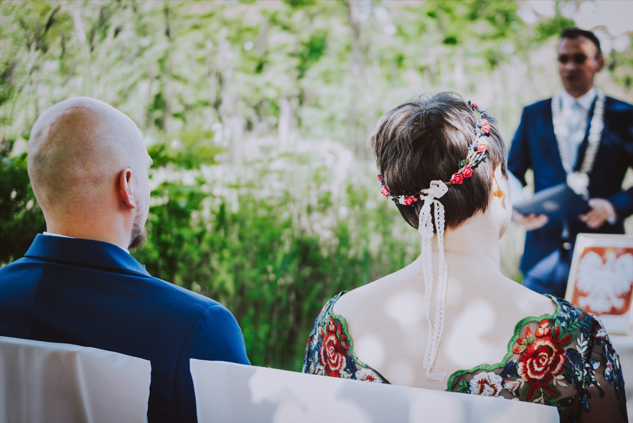 Para młoda siedzi podczas ceremonii ślubnej na tle zieleni, panna młoda w sukni z kwiatowym haftem i wiankiem na głowie, pan młody w granatowym garniturze, urzędnik w tle.