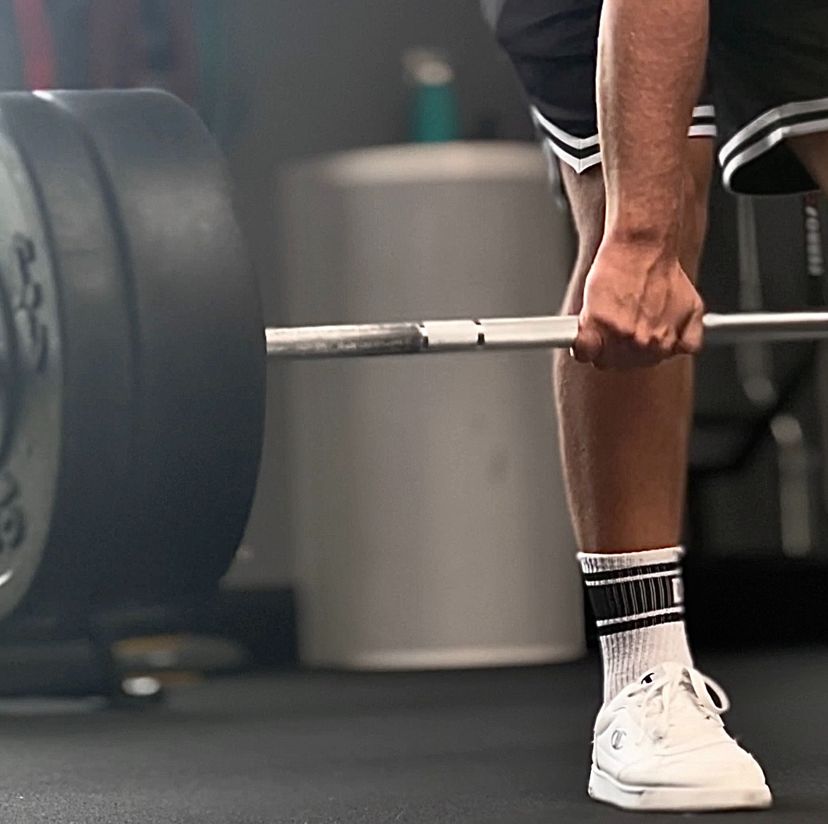 Mężczyzna w białych butach i czarno-białych skarpetkach przygotowuje się do podniesienia sztangi z obciążeniem w siłowni.