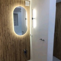 Jacek Sadowski - Nowoczesna łazienka z drewnianą okładziną ścienną, podświetlanym lustrem i minimalistycznym oświetleniem. Widoczne chromowane baterie i akcesoria.