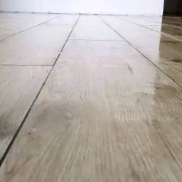 Jacek Sadowski - Błyszcząca, jasna podłoga wyłożona prostokątnymi płytkami imitującymi drewno, z widocznymi fugami. Minimalistyczne tło z białej ściany.