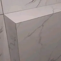 Białe płytki imitujące marmur, ułożone z precyzją na narożniku ściany. Widoczne fugi i delikatny wzór żyłkowania. Minimalistyczny design.