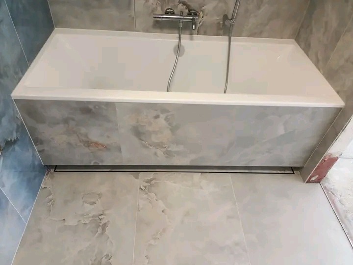 Wykończona wanna obudowana płytkami imitującymi marmur, z widocznym odpływem liniowym. Płytki podłogowe i ścienne w podobnej stylistyce.