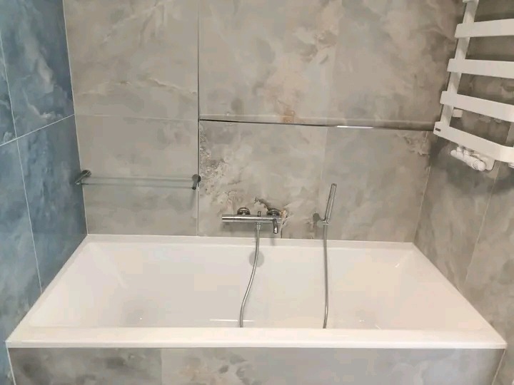 Biała wanna z chromowaną baterią na tle ściany z szarych płytek imitujących marmur. Widoczny grzejnik drabinkowy i szklana półka.